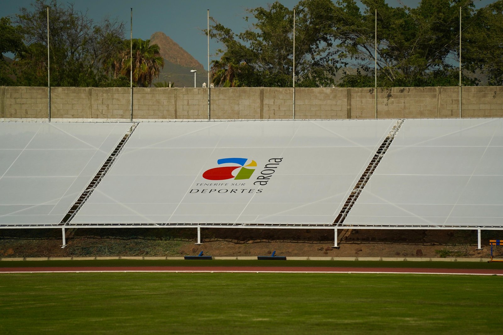 El Ayuntamiento de Arona ha concluido la instalación de lonas de gran formato en el Estadio Antonio Domínguez. Con una inversión de aproximadamente 56.000 euros, se han colocado estas lonas en la zona de la contra recta del estadio.
Además, se han iniciado las labores de mejora y acondicionamiento del césped natural del Estadio Antonio Domínguez, y se han finalizado las obras de insonorización y mejora del pabellón municipal de Arona casco.
Estas acciones forman parte de las estrategias destinadas a mejorar las instalaciones deportivas del municipio. El proyecto ha consistido en la colocación de ocho lonas microperforadas de 12 metros de ancho por 12 de largo, con un sistema de anclaje especializado que garantiza su resistencia y durabilidad.
Estas mejoras buscan embellecer y optimizar visualmente el espacio, utilizado por entidades deportivas locales y deportistas individuales y colectivos. El compromiso del Ayuntamiento con la modernización y el mantenimiento responsable de sus infraestructuras deportivas se refleja en estas intervenciones. Esta acción responde a la necesidad de mantener los estándares de calidad exigidos por federaciones deportivas y clubes usuarios del recinto, en el marco de la planificación continua realizada por el área de Deportes del Ayuntamiento para garantizar la excelencia y seguridad de las infraestructuras deportivas municipales.